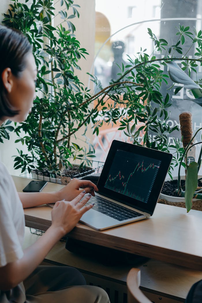 Businesswoman checks financial graphs on laptop in cozy café, surrounded by plants, showcasing remote work lifestyle.